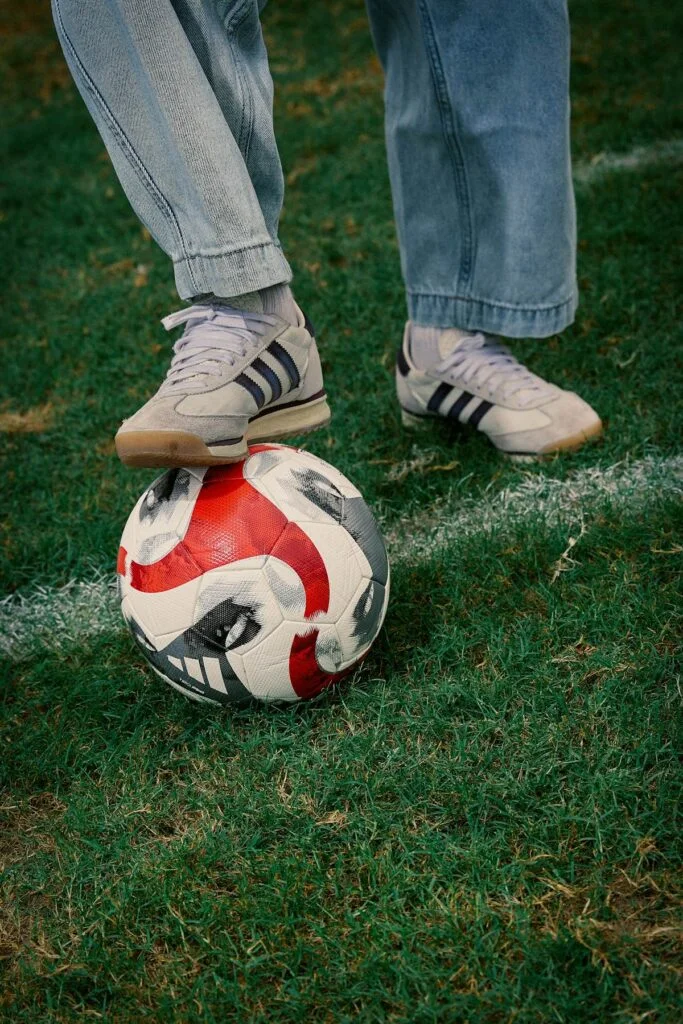 Feet in sneakers resting on a soccer ball on grass.